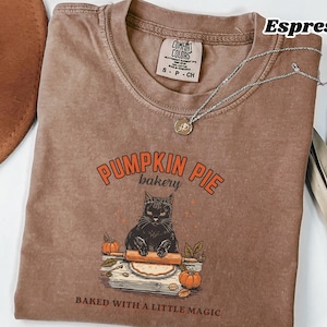 Puede incluir: Una camiseta color espresso con un gráfico de un gato negro usando un rodillo sobre masa. El texto dice PUMPKIN PIE bakery y BAKED WITH A LITTLE MAGIC. La camiseta tiene una etiqueta de Comfort Colors en el cuello y un collar plateado.