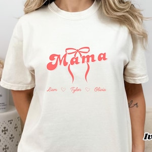 Op de afbeelding: Ivoor kleurig t-shirt met het woord "Mama" in een retro lettertype, versierd met een strik en vloeiende lijnen. De namen Liam, Tylor en Olivia staan eronder gedrukt. Een casual kledingstuk voor dagelijks gebruik.