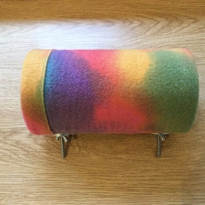 Puede incluir: Un tubo cubierto de forro polar de colores con un patrón de tie-dye en tonos de amarillo, naranja, rojo, morado y verde. El tubo está sujeto a una superficie de madera con dos ganchos metálicos.