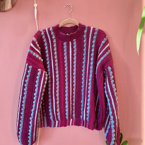 Könnte beinhalten: Ein weinroter Pullover mit vertikalen Streifen aus weißen und hellblauen Bommeln. Der Pullover hat einen Rundhalsausschnitt und lange Ärmel. Der Pullover hängt an einem Holzbügel vor einer rosa Wand.