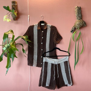 Könnte beinhalten: Ein braunes und weißes Häkel-Shirt und -Shorts-Set. Das Shirt hat einen Knopfleisten-Verschluss und kurze Ärmel. Die Shorts haben einen elastischen Bund und einen Kordelzug.