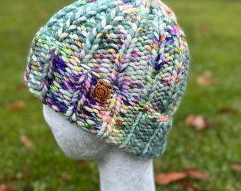 May include: A hand-knitted beanie hat in shades of green, blue, and multicolored yarn. The hat features a ribbed design and a small, brown leather tag with a smiling face. The hat is displayed on a mannequin head against a blurred green background.