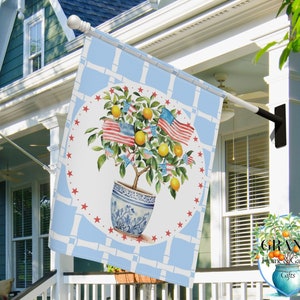 May include: A blue and white garden flag with a red, white, and blue American flag tree with lemons and stars. The flag is hanging on a white pole in front of a house.
