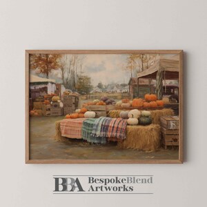 Op de afbeelding: Een ingelijste prent van een herfst oogsttafereel. Het kunstwerk toont een pompoenveld met hooibalen, houten kisten en een verscheidenheid aan pompoenen in oranje, wit en groen. De scène speelt zich af in een landelijk landschap met bomen en gebouwen.