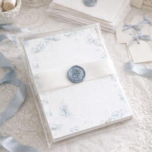 May include: A stack of rectangular stationery cards featuring a light blue floral border pattern, enclosed in a clear acrylic case. A white semi-transparent ribbon wraps around the center, secured by a circular blue wax seal with an embossed design.