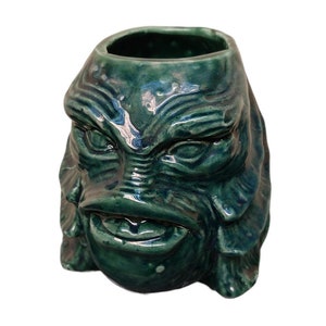 May include: Green ceramic mug shaped like the Creature from the Black Lagoon. The mug has a detailed face with gills, eyes, and a mouth.