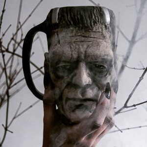 May include: A ceramic mug shaped like Frankenstein's head. The mug is gray and has a textured surface. The mug has a handle on the side.