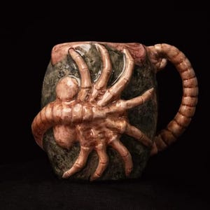 May include: A ceramic mug shaped like an alien creature bursting out of an egg. The mug has a brown and green exterior with a detailed alien design. The handle is shaped like the alien's tail.