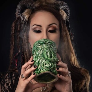 May include: A woman with long brown hair and horns drinks from a green ceramic mug shaped like a Cthulhu monster.