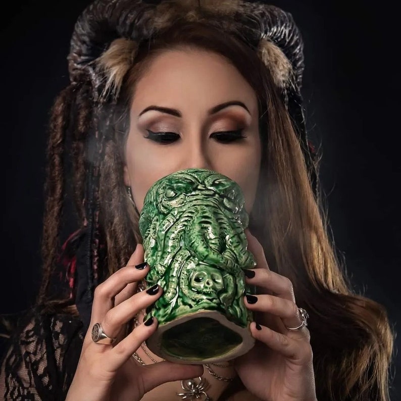 May include: A woman with long brown hair and horns drinks from a green ceramic mug shaped like a Cthulhu monster.