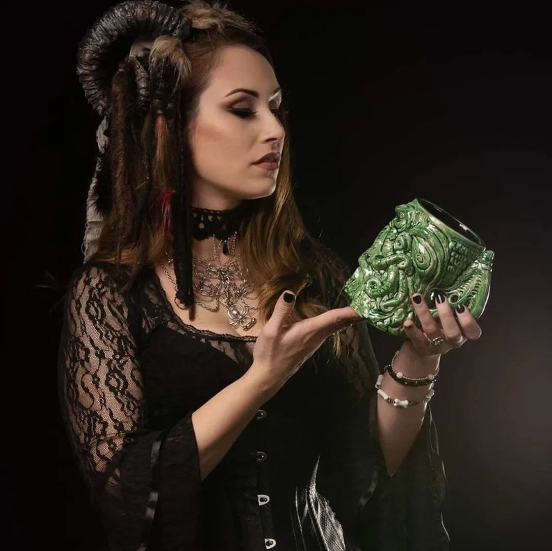 May include: A woman with dark hair and horns is holding a green ceramic mug shaped like a monster's head.