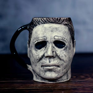 Op de afbeelding: Een keramische mok in de vorm van het hoofd van Michael Myers uit de Halloween-horrorfilmfranchise. De mok heeft een zwart handvat en een wit, gedetailleerd gezicht met zwarte ogen.
