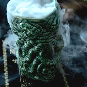 May include: Green ceramic mug shaped like a monster's head, filled with white smoke. The mug is sitting on a black surface with gold lettering that reads "Eternal Heavens".