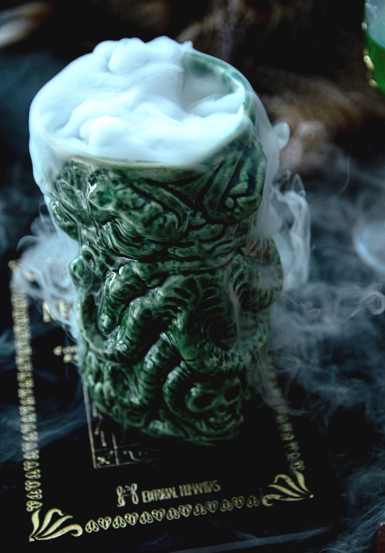 May include: Green ceramic mug shaped like a monster's head, filled with white smoke. The mug is sitting on a black surface with gold lettering that reads "Eternal Heavens".