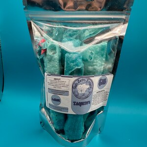 以下が含まれることがあります： 青い円筒形のトリートが入った透明なビニール袋。袋には「Sub Zero Treats」と「TAQUITO'S」の文字が入ったラベルが付いています。トリートはフリーズドライのようです。背景は単色の水色です。