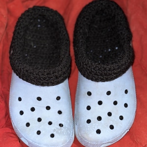 May include: A pair of light blue clogs with black crocheted cuffs. The clogs have a textured sole with small holes.
