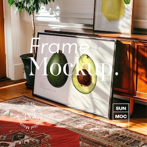 May include: A black framed print of two avocado halves, one whole and one sliced open, with the text "Frame Mockup." on a white background. The print is sitting on a wooden table with a rug in front of it.