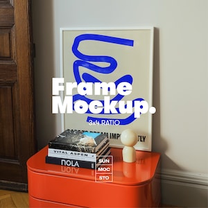 May include: A framed art print with a white border featuring blue abstract shapes on a beige background. The text "Frame Mockup" is displayed. The frame rests on a bright orange nightstand with books and a small wooden figurine.