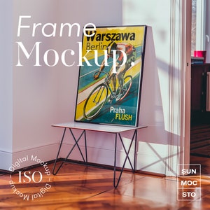 Puede incluir: Un póster enmarcado en negro con una ilustración de ciclismo vintage. El póster presenta a un ciclista con un maillot amarillo y azul montando una bicicleta en una pista. El póster está titulado "Warszawa Berlin Praha FLUSH". El póster está sentado en una mesa de metal negro en una habitación con suelo de madera.