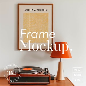 Puede incluir: Una impresión enmarcada con el texto "WILLIAM MORRIS" y un patrón floral amarillo. El marco es de madera marrón. Un tocadiscos con una plataforma giratoria negra y una lámpara roja con una base beige están sobre una superficie de madera. El texto "Frame Mockup." está en letras blancas sobre un fondo blanco.