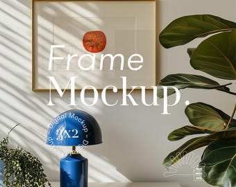 May include: A gold picture frame mockup with a blue lamp on a white table. The frame is on a white wall with a green plant in the background.