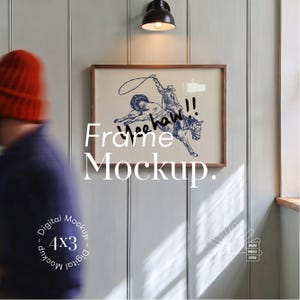 May include: A rectangular wooden frame containing a print of a cowboy riding a bucking horse with a lasso, featuring the text Yeehaw!! in blue ink. The frame is mounted on a light green paneled wall beneath a black wall-mounted light fixture.