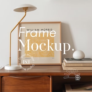 以下が含まれることがあります： 白いシェードのついた金色調のテーブルランプが、黄色と白のパターンが描かれた額縁付きのプリントが置かれた木製デスクの上に置かれています。プリントには「WILLIAM MORRIS Frame Mockup.」というテキストが書かれています。デスクには引き出しがあり、本と白いセラミック製のオブジェクトが置かれています。