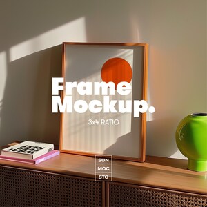 May include: A framed art print with an orange circle design, labeled "Frame Mockup" with a 3x4 ratio. The frame is displayed on a wooden cabinet with books and a green vase. The image is lit by sunlight.