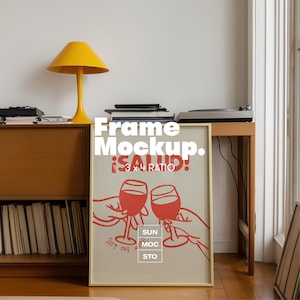 May include: A framed poster mockup with a beige background and red graphic of two wine glasses clinking. The poster has the text "Frame Mockup. ¡SALUD! 3 x 4 RATIO" and "JUST ONE MOC STO". A yellow lamp and a wooden cabinet are in the background.