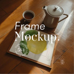 May include: A wooden frame mockup with a digital mockup text overlay. The frame is holding a print of a lemon with green leaves. The frame is sitting on a wooden surface with a teacup and teapot in the background.
