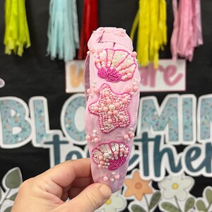 May include: A pink headband adorned with beaded seashell and starfish designs. The headband features a variety of pink beads and sequins. The background includes colorful tassels and the words "BLOOMING TOGETHER".