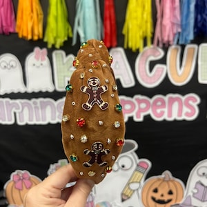 May include: A brown velvet headband adorned with gingerbread man charms and colorful rhinestones. The headband is embellished with clear and colored gems. The background features colorful tassels and the words "MAGICAL LEARNING HAPPENS".