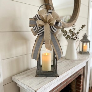 May include: A metal lantern featuring a large decorative bow made of tan burlap and black and white striped ribbon. Inside the lantern sits a single cream-colored pillar candle with a warm glowing light, creating a soft illumination within the frame.