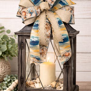 May include: A dark gray wooden lantern with a lit candle inside, decorated with seashells and a large bow. The bow features a beach-themed pattern with blue, yellow, and beige colors. The lantern is a decorative home accent.