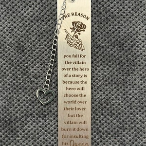 May include: A silver bookmark with a black rose and skeleton hand design. The bookmark has a chain and a heart charm. The text on the bookmark reads "THE REASON you fall for the villain over the hero of a story is because the hero will choose the world over their lover but the villain will burn it down for insulting his Queen."