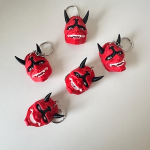 May include: Red devil mask keyrings with black horns, eyes, and facial details. The keyrings have a silver ring and chain. The masks have a smiling expression with white teeth. The keyrings are arranged on a white surface.