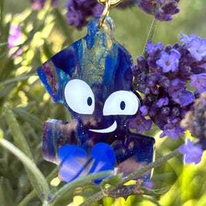 May include: A puzzle piece keychain featuring a swirling dark blue, purple, and gold glitter resin finish. The piece has a cartoon face with two large white eyes and a small white mouth, plus two solid blue feet at the bottom, attached to a gold ring.