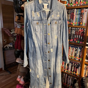 May include: A light blue denim duster jacket with distressed detailing, featuring button closures and pockets. The longline coat is hanging on a hanger. The jacket is a casual outerwear garment.