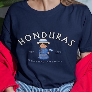 May include: A navy blue t-shirt with a white graphic of a girl wearing a traditional Honduran dress and holding a flag. The text "HONDURAS" is in a white arc above the girl, and the text "EST. 1821 CENTRAL AMERICA" is below her.