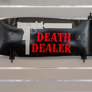 May include: Black skateboard deck with a white silhouette of a gun and the words "DEATH DEALER" in red lettering.
