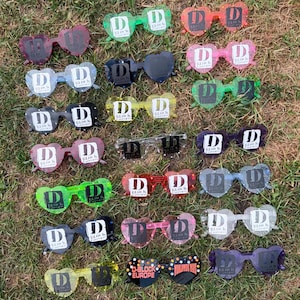 May include: A collection of heart-shaped sunglasses in various colours including red, green, blue, yellow, pink, black, and clear. Each pair features the D-Block Europe logo printed on the lenses. The frames are made of a translucent material.