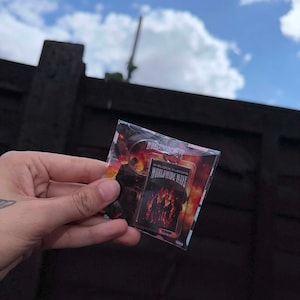 May include: A rectangular metal lighter featuring a graphic print of the O-Block Europe Worldwide Wave album cover, which depicts a fiery orange and red flame background with silhouettes of figures, all encased in a clear plastic protective sleeve.