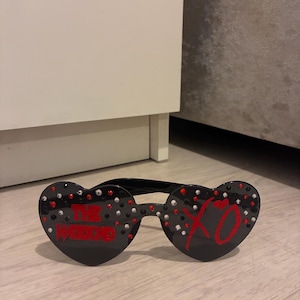 May include: Black heart-shaped sunglasses with the text "THE WEEKND" in red on one lens and "XO" on the other. The sunglasses are decorated with red and silver rhinestones.