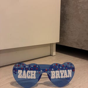 Puede incluir: Gafas de sol azules en forma de corazón con el texto "ZACH BRYAN" en letras blancas. Las gafas están decoradas con pedrería roja y plateada. Un accesorio divertido.