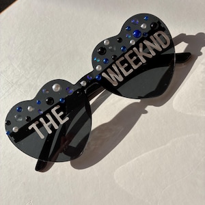 May include: Black heart-shaped sunglasses with the text "THE WEEKND" in silver. The sunglasses are decorated with small, colourful rhinestones. The lenses are dark, and the frames are black.