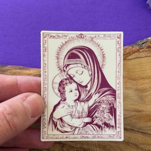 May include: A sticker with a vintage illustration of the Virgin Mary holding baby Jesus. The image is in shades of pink and white on a cream background.