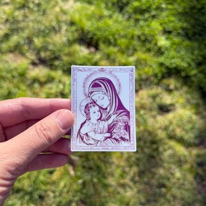May include: A rectangular sticker featuring a religious image of a woman and child in shades of purple and white. The woman wears a veil and holds the child, both with halos. The image is framed with a decorative border.