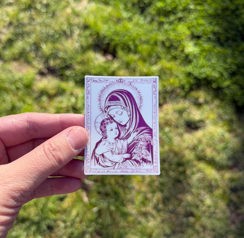 May include: A rectangular sticker featuring a religious image of a woman and child in shades of purple and white. The woman wears a veil and holds the child, both with halos. The image is framed with a decorative border.