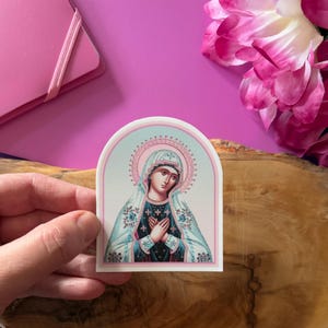 May include: A sticker featuring a religious icon of the Virgin Mary. The image is in a traditional style with a blue and white robe and a pink halo. The sticker is on a white background with a rounded arch shape.