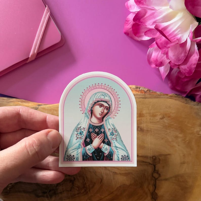 May include: A sticker featuring a religious icon of the Virgin Mary. The image is in a traditional style with a blue and white robe and a pink halo. The sticker is on a white background with a rounded arch shape.